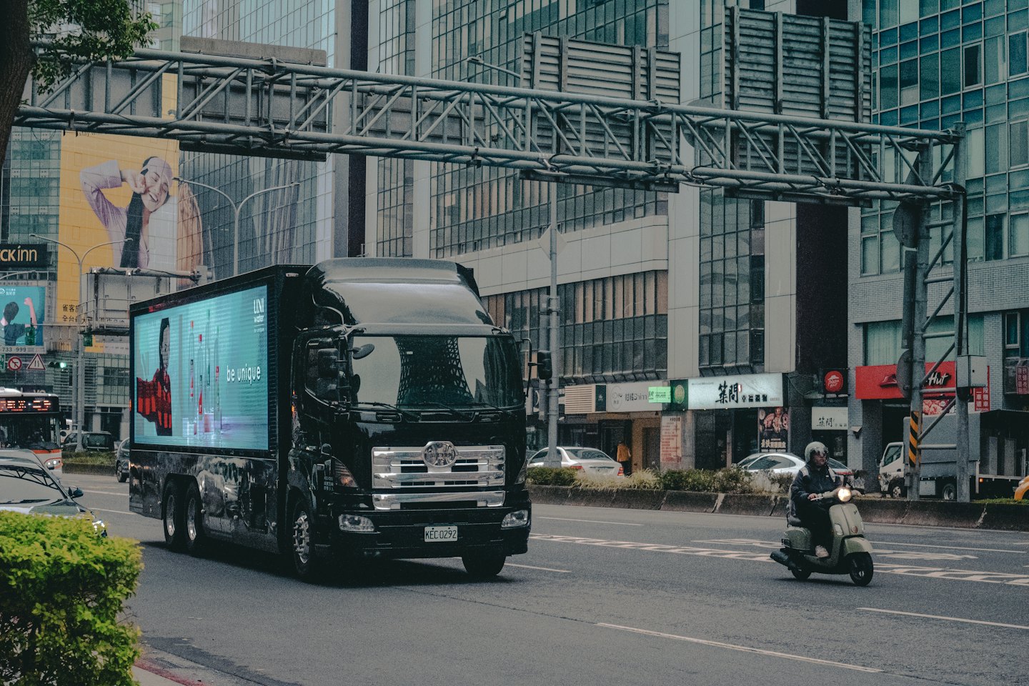 A digital billboard truck drives past city buildings and a scooter.