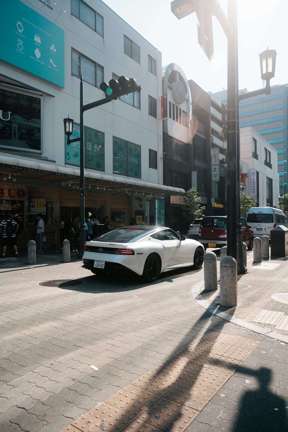 White sports car drives down a sunlit city street past storefronts.